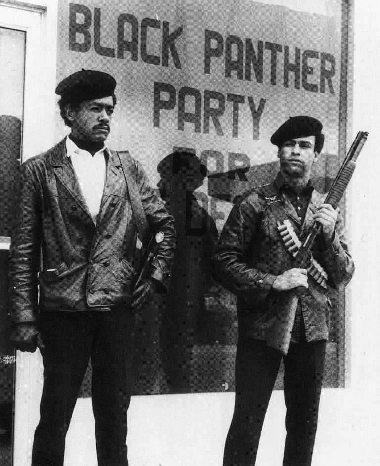 Bobby Seale and Huey Newton stand unsmiling in front of Black Panther Party Banner